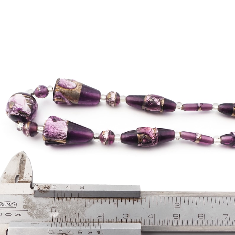 May include: A necklace featuring a series of purple glass beads with metallic accents. The beads vary in shape, including round and elongated designs, strung together with clear beads. A silver measuring tool is in the foreground.