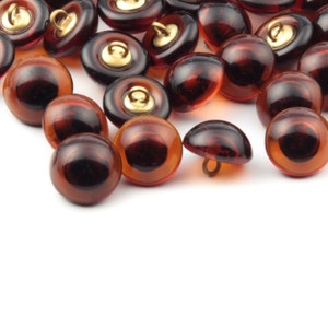 May include: A collection of brown glass buttons with gold accents. The buttons are arranged in a random pattern on a white surface.