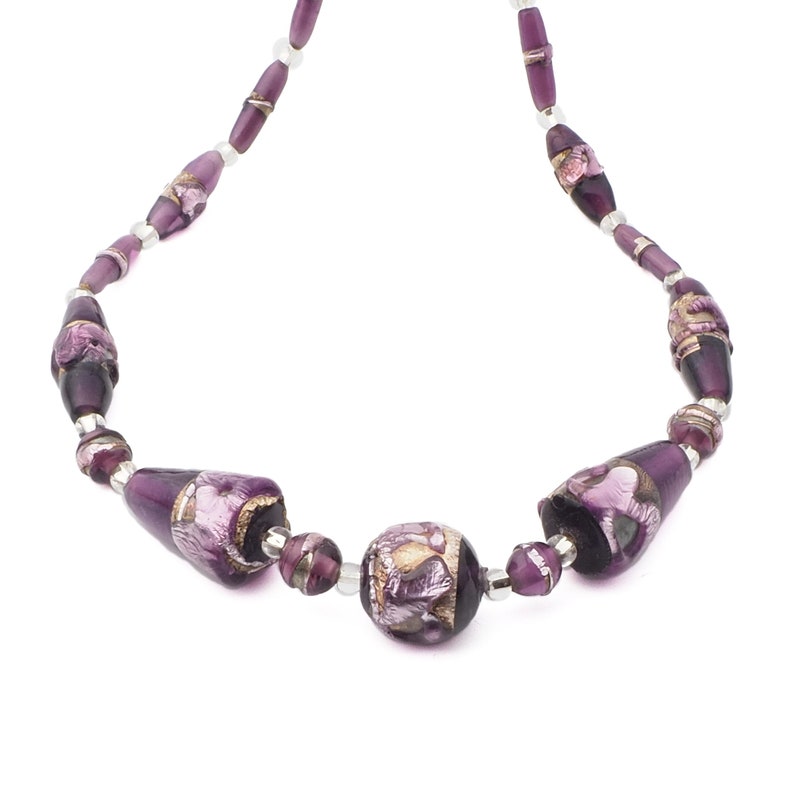 May include: A purple beaded necklace with a mix of shapes and sizes. The necklace features large, barrel-shaped beads and a central round bead, all in shades of purple with gold accents. Small silver-colored beads connect the larger beads.