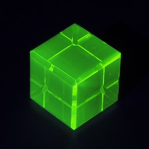 Può includere: Un cubo verde neon brillante con un design geometrico. Il cubo è realizzato in un materiale traslucido e presenta un motivo a griglia sulla superficie. L'oggetto è posto su uno sfondo nero.