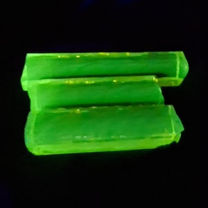 May include: Three bright green, rectangular objects are stacked against a black background. The objects have a slightly translucent appearance, with a few yellow specks visible within. The edges are angled, and the overall effect is a glowing, neon-like appearance.