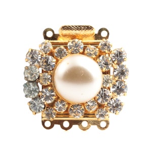 Puede incluir: Un cierre dorado con una perla blanca grande rodeada de diamantes de imitación transparentes.