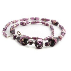 Puede incluir: Un collar artesanal con una variedad de cuentas moradas. El collar incluye cuentas alargadas en forma de barril, cuentas redondas m&aacute;s peque&ntilde;as y cuentas m&aacute;s grandes de forma irregular con detalles dorados. El collar est&aacute; ensartado en un cord&oacute;n transparente y flexible y tiene un cierre plateado.
