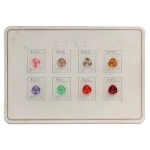 May include: A vintage catalog page displaying eight triangular glass beads in various colours. The beads are pink, orange, green, red, and blue. Each bead is labelled with a number and other handwritten markings on the aged paper.