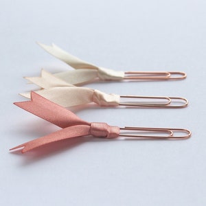 May include: Three gold paper clips with pink and white satin ribbon bows. The bows are tied in a knot and the ends of the ribbon are fanned out.