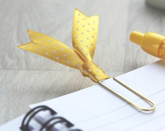 May include: A yellow polka dot ribbon bookmark attached to a gold paper clip on a white spiral bound notebook.