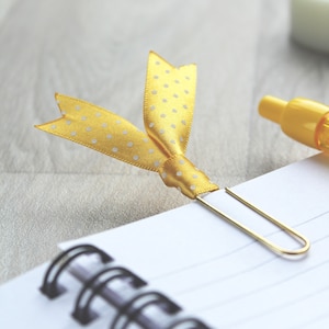 May include: A yellow polka dot ribbon bookmark attached to a gold paper clip on a white spiral bound notebook.