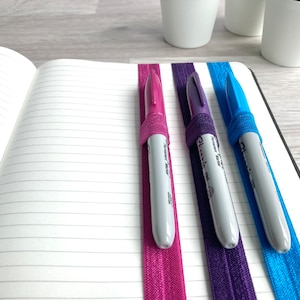 May include: Three pens are held in place by elastic bands in a notebook. The bands are pink, purple, and blue. The pens are grey and have black caps.