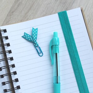 May include: A blue spiral bound notebook with a turquoise ribbon bookmark and a turquoise pen. The notebook has white lined paper.