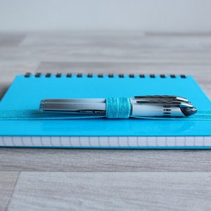 May include: A blue spiral bound notebook with a silver and black pen on top. The pen is wrapped in a blue ribbon.