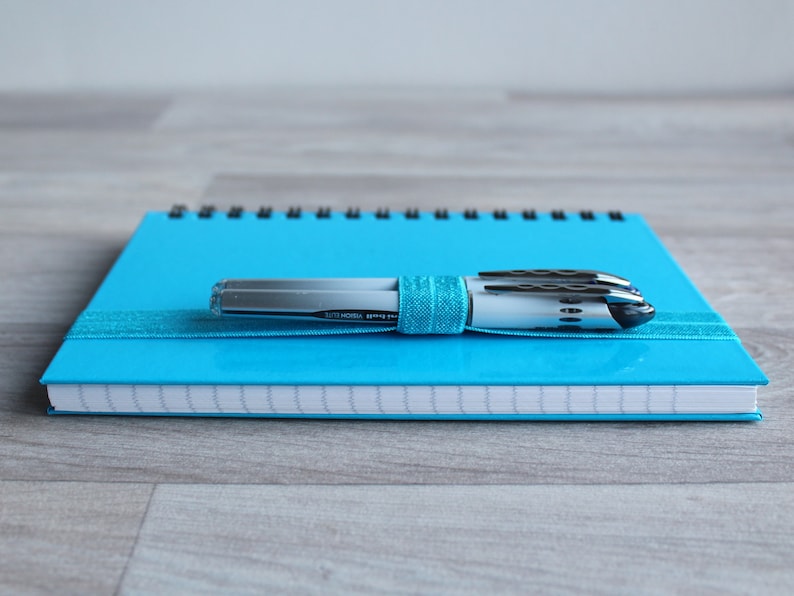May include: A blue spiral bound notebook with a silver and black pen on top. The pen is wrapped in a blue ribbon.