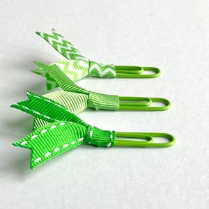 May include: Three green paper clips with green and white ribbon bows. The bows are tied in a knot and the ends are fanned out.