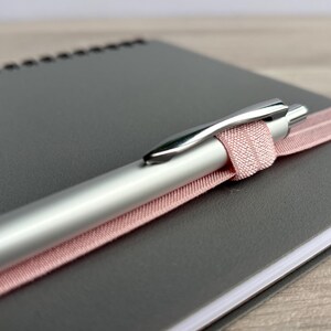 Puede incluir: Un bol&iacute;grafo plateado con una cinta rosa envuelta alrededor, descansando sobre un cuaderno gris.