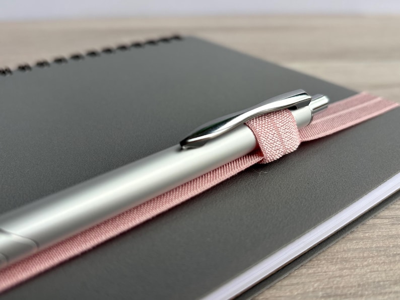 Puede incluir: Un bol&iacute;grafo plateado con una cinta rosa envuelta alrededor, descansando sobre un cuaderno gris.