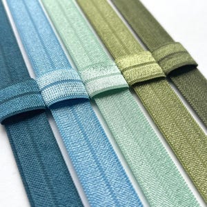 May include: Close-up of six elastic hair ties in shades of blue and green. Each tie has a textured band and a folded-over section. The colours range from teal to olive green, creating a gradient effect. The background is white.