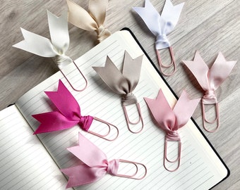May include: Six pink and white ribbon bookmark clips. The clips are made of metal and have a decorative ribbon tied around them. The ribbons are in various shades of pink and white.