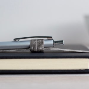 May include: A silver pen with a gray fabric pen holder is resting on a black and cream notebook.