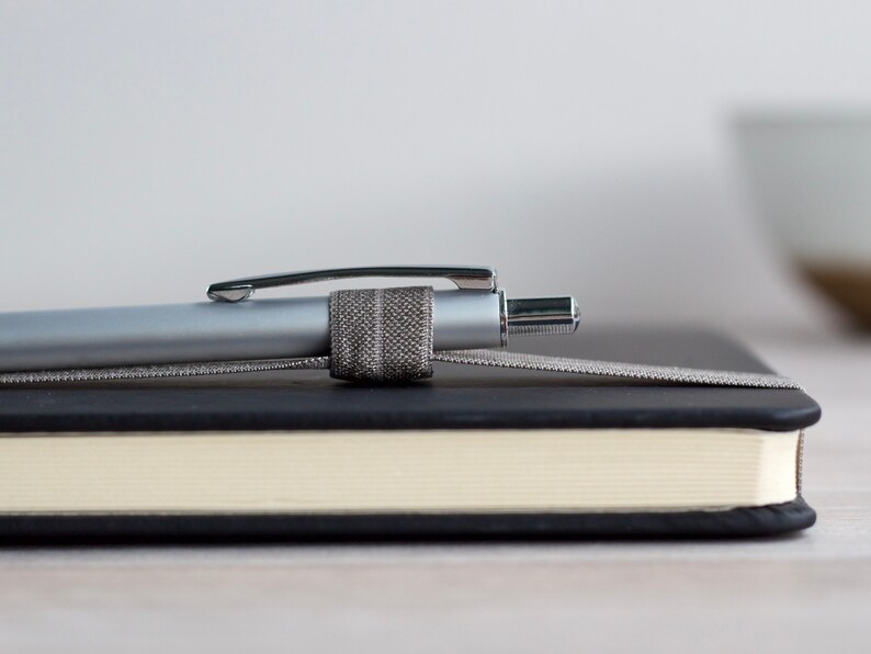 May include: A silver pen with a gray fabric pen holder is resting on a black and cream notebook.