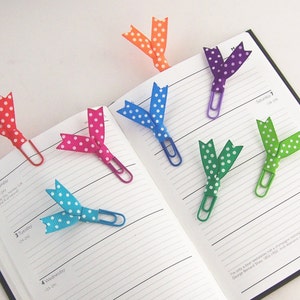 May include: A collection of colorful polka dot ribbon bookmarks attached to paper clips, resting on the open pages of a planner. The ribbons are in various colors, including red, pink, blue, green, purple, and orange.