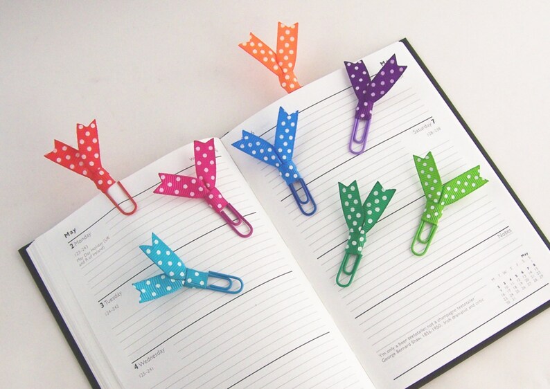 May include: A collection of colorful polka dot ribbon bookmarks attached to paper clips, resting on the open pages of a planner. The ribbons are in various colors, including red, pink, blue, green, purple, and orange.