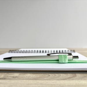 Puede incluir: Un bol&iacute;grafo plateado con una banda el&aacute;stica verde descansa sobre un cuaderno blanco con una encuadernaci&oacute;n en espiral negra.