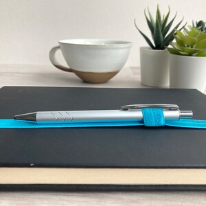 May include: A black notebook with a silver pen and a blue elastic band. The pen is resting on the notebook.