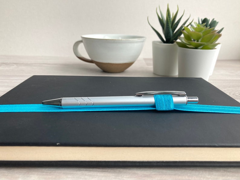 May include: A black notebook with a silver pen and a blue elastic band. The pen is resting on the notebook.