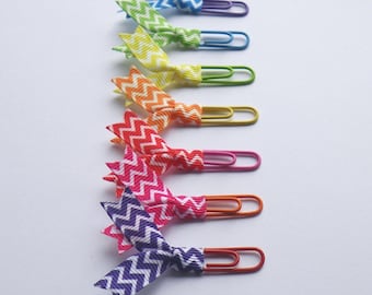 May include: A set of seven colorful paper clips with rainbow chevron patterned ribbon bows. The paper clips are arranged vertically in a rainbow order from blue to purple.