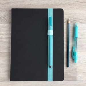 May include: A black notebook with a blue ribbon bookmark and a blue pen attached to it. A pencil and a blue pen are lying on a wooden surface next to the notebook.
