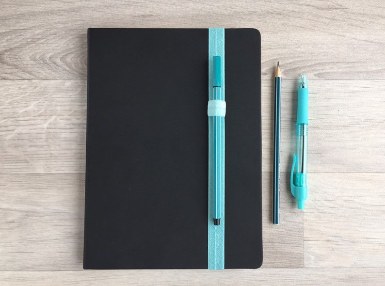 May include: A black notebook with a blue ribbon bookmark and a blue pen attached to it. A pencil and a blue pen are lying on a wooden surface next to the notebook.