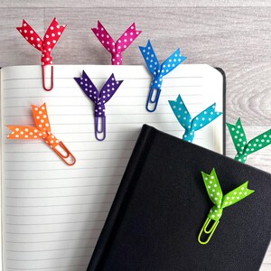 May include: A collection of colorful polka-dot ribbon bookmarks attached to paperclips, resting on the pages of an open notebook and a closed black book. The ribbons are red, pink, blue, orange, purple, teal, and green.