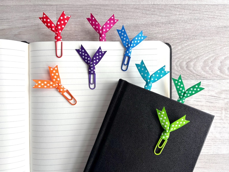 May include: A collection of colorful polka-dot ribbon bookmarks attached to paperclips, resting on the pages of an open notebook and a closed black book. The ribbons are red, pink, blue, orange, purple, teal, and green.