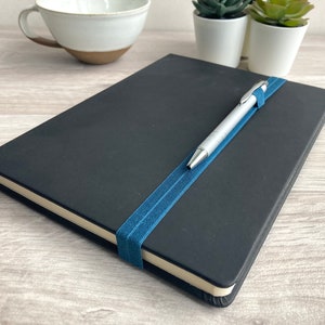 May include: A black notebook with a blue elastic band holding a silver pen in place.