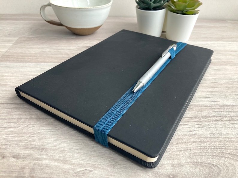 May include: A black notebook with a blue elastic band holding a silver pen in place.
