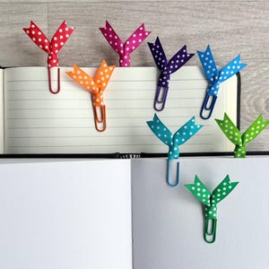 May include: A collection of colorful polka-dot ribbon bookmarks attached to paperclips, inserted into the pages of an open notebook. The ribbons are red, pink, orange, purple, blue, teal, and green. The notebook has lined pages.