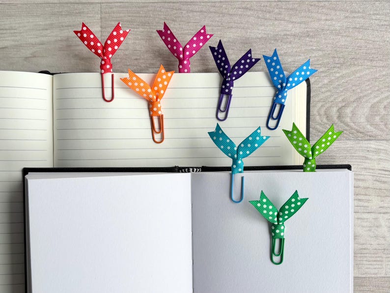 May include: A collection of colorful polka-dot ribbon bookmarks attached to paperclips, inserted into the pages of an open notebook. The ribbons are red, pink, orange, purple, blue, teal, and green. The notebook has lined pages.