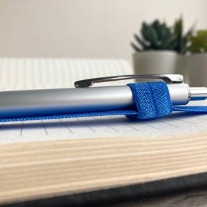 May include: A silver pen with a blue elastic band is lying on a lined notebook. The notebook is closed and has a brown cover.