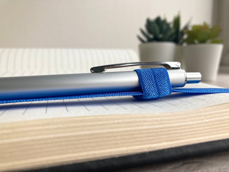 May include: A silver pen with a blue elastic band is lying on a lined notebook. The notebook is closed and has a brown cover.