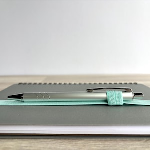Puede incluir: Un cuaderno gris de espiral con un bol&iacute;grafo plateado y una banda el&aacute;stica azul claro que sujeta el bol&iacute;grafo en su lugar.