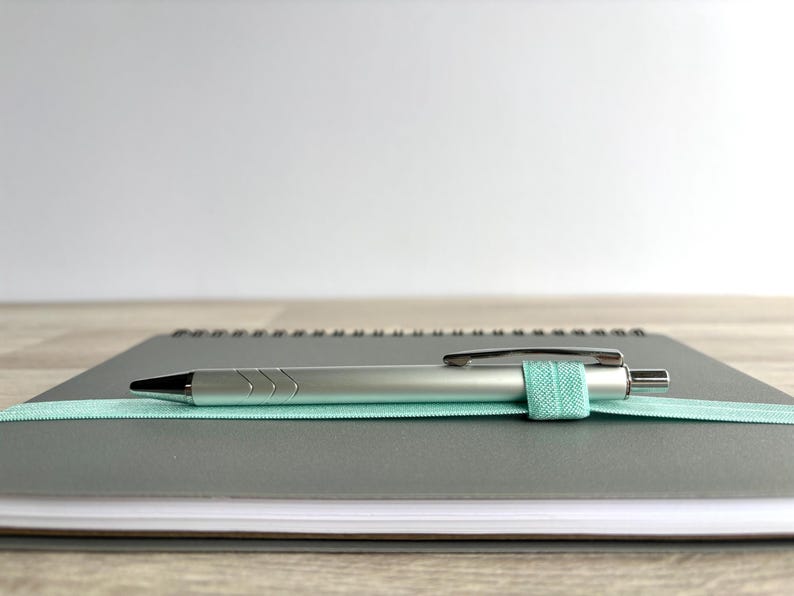 Puede incluir: Un cuaderno gris de espiral con un bol&iacute;grafo plateado y una banda el&aacute;stica azul claro que sujeta el bol&iacute;grafo en su lugar.