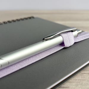 Puede incluir: Un bol&iacute;grafo plateado con una cinta lavanda envuelta alrededor, descansando sobre un cuaderno gris.