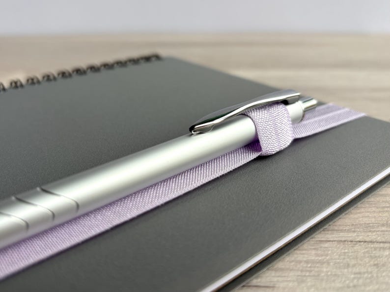 Puede incluir: Un bol&iacute;grafo plateado con una cinta lavanda envuelta alrededor, descansando sobre un cuaderno gris.