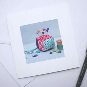May include: A colourful fabric pincushion in the shape of a cube with colourful pins sticking out of the top. The pincushion is surrounded by buttons, a spool of thread, and a pair of scissors.