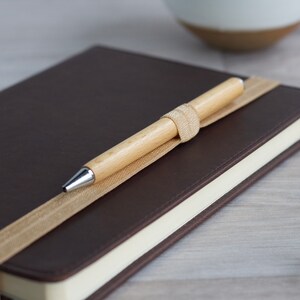 May include: A brown leather-bound journal with a beige ribbon bookmark and a wooden pen with a silver tip.