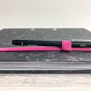 May include: A black binder with a pink elastic band holding a black pen with the text "0.5 mm ROTTER & KRONE" on it.