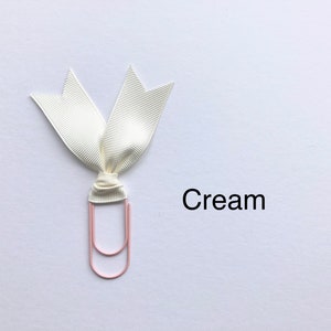 Może przedstawiać: Wstążka w kolorze kremowym zawiązana wok&oacute;ł r&oacute;żowego spinacza do papieru. Tekst "Krem" jest napisany pod wstążką.