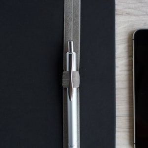 May include: A silver pen is held in place by a gray elastic band on a black notebook.