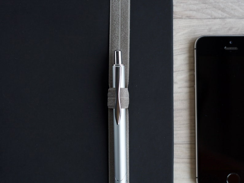 May include: A silver pen is held in place by a gray elastic band on a black notebook.