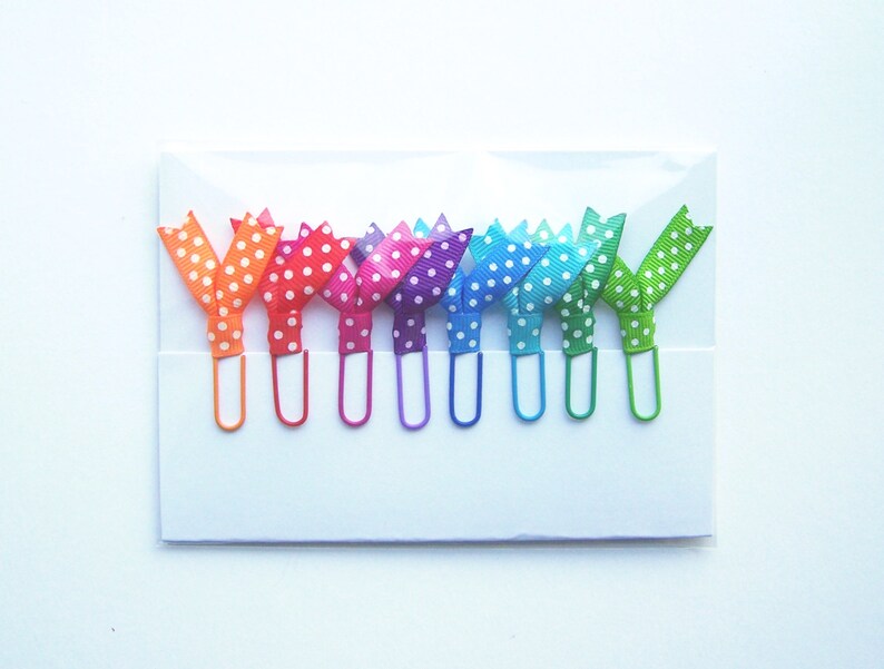 May include: A set of colorful paper clips with polka dot ribbon bows. The clips are in a variety of colors, including orange, red, pink, purple, blue, green, and yellow. Each clip is attached to a white card.