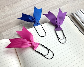 May include: Three colorful ribbon bookmarks with black metal clips. The bookmarks are pink, purple, and blue.